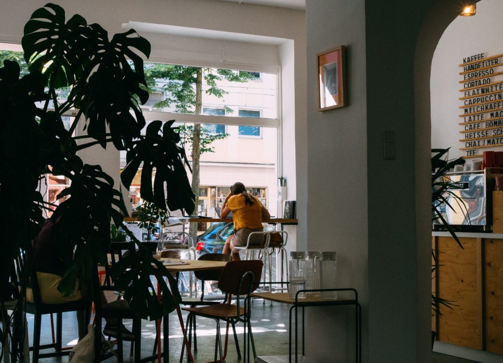 Digital nomad wearing a mustard top and ochre shorts working from a laptop at a café. Sitting by a bright window facing the street. 