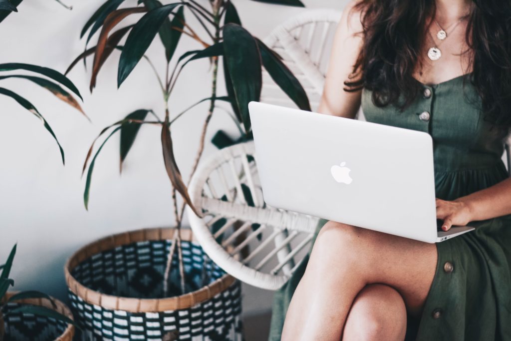 Young woman with long dark brown hair wearing a dark green dress working with a laptop on her lap. She's sitting on a white chair and in the background there are some plants in front of a white wall.