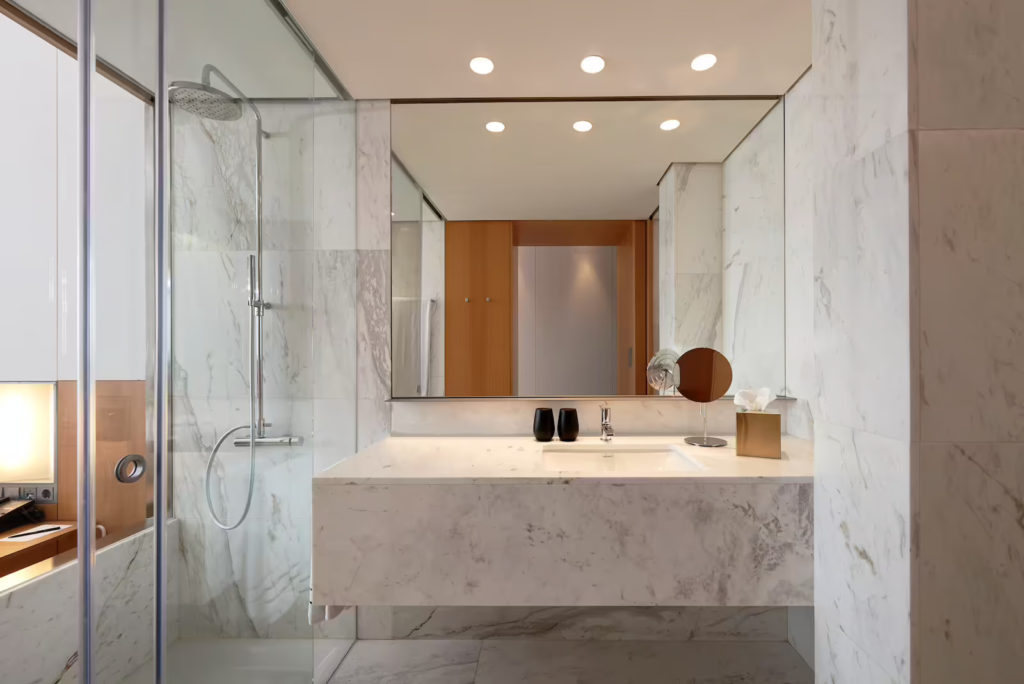 Image of a white marble bathroom. It faces the sink with a mirror above it and a glass shower panel on the left.