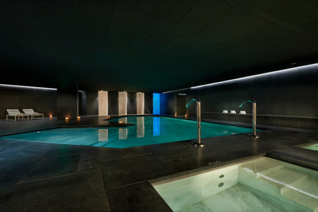 Image of a swimming pool with dark grey tiles surrounding it. There are two white lounges in the background on the left and four shower areas in front.