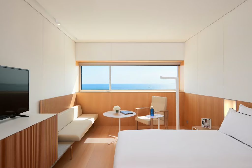 View of a room with the bed on the right-hand side, a black TV on top of a cabinet on the left. In the background, there's a   panoramic window from where you can see the ocean, a white leather seat on the left, a chair of the same material on the right and a round, wooden table in the middle. 