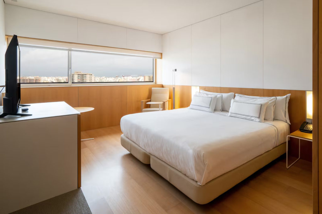 Photo of the room at an angle, facing the right back corner. The bed covered with white linen and a bedside table is on the right-hand side. A black TV is on top of a cabinet on the left hand-side. In the background, a round wooden table is partially shown immediately behind the cabinet and there's a wooden chair with cream leather seat in the right back corner. The room is bright and it has a sea view.