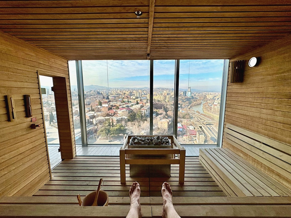This photo shows the sauna's wood-panelled interior, with a water bucket next to Paul's feet and a glass door to his left, while there's a large square recipient storing sauna rocks in the middle as well as a rectangular window overlooking the city in front.