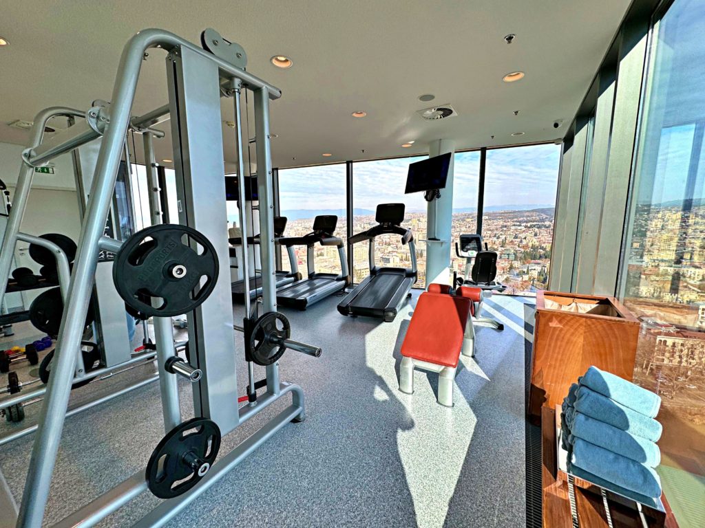 View of the spa fitness room framed by glass windows, offering city views. In the foreground, a weight machine on the left, rolled blue towels on a wooden rack on the right, and a brown leather exercise bench in between. In the background, three black treadmills align between two white columns, each with a black TV screen.