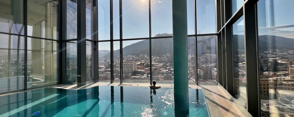 View of the spa pool with sunlight streaming through the windows, reflecting the window dividers on the azure swimming pool. In the distance, Milene can be seen in the pool, facing away from the camera and enjoying the views through the glass windows of the spa.