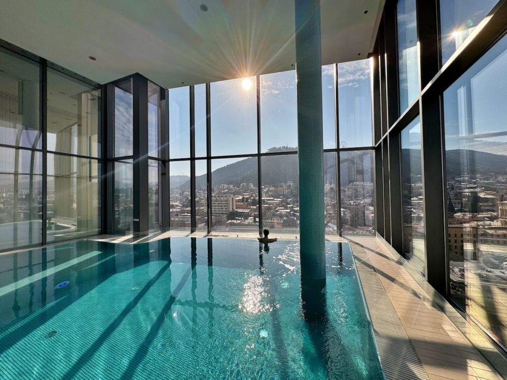 View of the spa pool with sunlight streaming through the windows, reflecting the window dividers on the azure swimming pool. In the distance, Milene can be seen in the pool, facing away from the camera and enjoying the views through the glass windows of the spa.