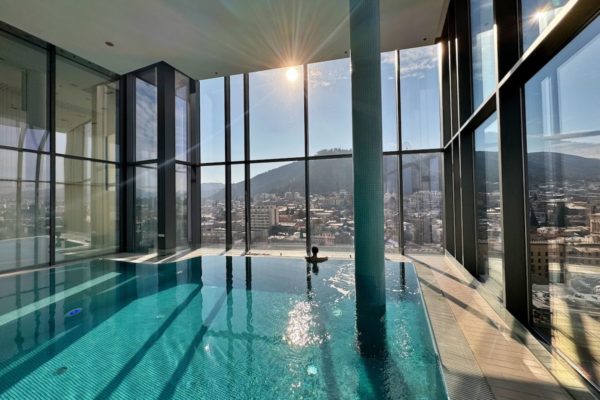 View of the spa pool with sunlight streaming through the windows, reflecting the window dividers on the azure swimming pool. In the distance, Milene can be seen in the pool, facing away from the camera and enjoying the views through the glass windows of the spa.