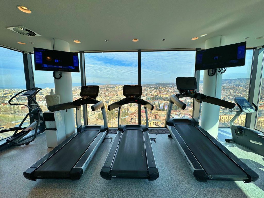 Three black treadmills aligned between two white columns, each featuring a black TV screen, facing the window with scenic views of Tbilisi.