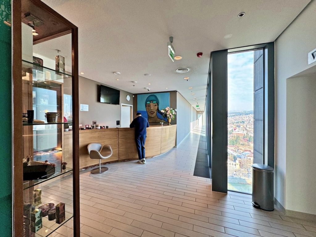 In the foreground on the left, a glass cupboard with wooden framing stores spa products. In the background, a man wearing a navy top, trousers, and white trainers stands in front of the spa's curved wood reception desk. The walls on the left are light brown, adorned with a TV screen, a white door, a vase with white and pink flowers, and a painting featuring a swimmer in a blue cap and goggles against a serene ocean and sky backdrop. To the right, a narrow window reveals city views and a long corridor leading to the spa.