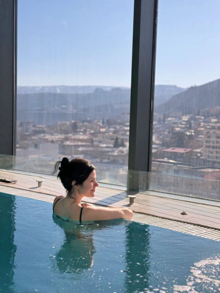 Milene is in the pool, wearing a black bikini with her hair in a bun, facing away from the camera and gazing through the glass windows of the spa, enjoying scenic views of Tbilisi.