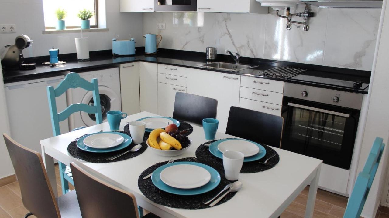 A white furnished kitchen with a rectangular dining table in the middle with six chairs of different colours and shapes around it. On the table there are four sets of white plates on top of blue plates and black placemats.