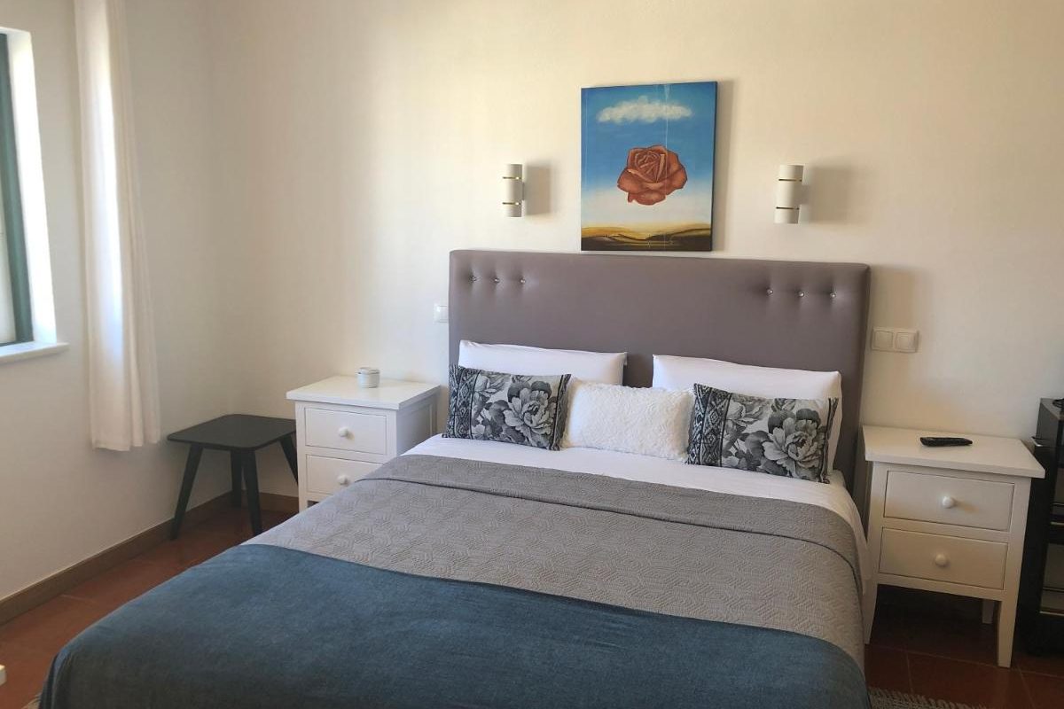 Image of a double bed with a grey headboard and two white bedside tables. There's also a picture on the wall above the bed.