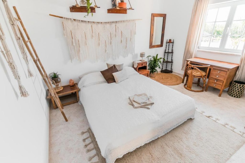 Room at the Lemontree Eco Surfhouse in Sagres predominantly white. There are some wooden furniture such as bedside tables and a desk and chair next to the bed on the right in front of a window.