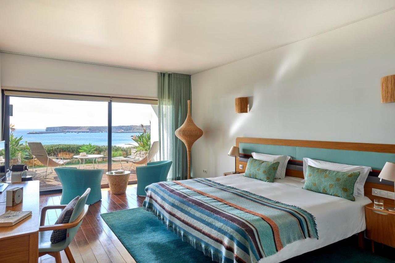 Room at Martinhal Sagres Beach Resort decorated with a combination of white, blue and turquoise tones. There's a wooden desk with a chair in front and two round chairs in the background. Both chairs face a window with a view of the ocean.