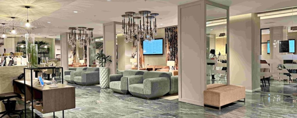 In the image, the hotel reception area is depicted. The walls and ceiling are painted in a light pinkish-grey colour, while the floor is adorned with grey marble tiles,. On the left side of the image, there is the reception desk and on the right side, there are three columns with mirrors attached from top to bottom on the side that faces the camera. Between the columns, there are two velvety grey sofas and a plant in a white vase is placed in between the sofas. In the background, there are additional seating areas with pink couches. To illuminate the reception area, there are several glass ceiling pendant lights hanging from above. The ones above the reception desk are shaped like upside-down umbrellas, while the ones above the sofas are modern grouped round balls covering the bulbs.