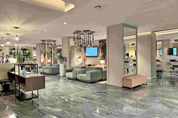 In the image, the hotel reception area is depicted. The walls and ceiling are painted in a light pinkish-grey colour, while the floor is adorned with grey marble tiles,. On the left side of the image, there is the reception desk and on the right side, there are three columns with mirrors attached from top to bottom on the side that faces the camera. Between the columns, there are two velvety grey sofas and a plant in a white vase is placed in between the sofas. In the background, there are additional seating areas with pink couches. To illuminate the reception area, there are several glass ceiling pendant lights hanging from above. The ones above the reception desk are shaped like upside-down umbrellas, while the ones above the sofas are modern grouped round balls covering the bulbs.