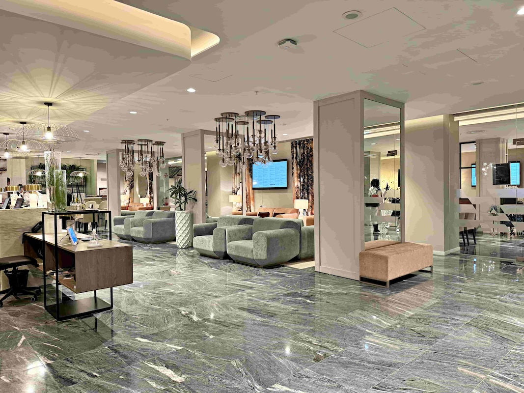 In the image, the hotel reception area is depicted. The walls and ceiling are painted in a light pinkish-grey colour, while the floor is adorned with grey marble tiles,. On the left side of the image, there is the reception desk and on the right side, there are three columns with mirrors attached from top to bottom on the side that faces the camera. Between the columns, there are two velvety grey sofas and a plant in a white vase is placed in between the sofas. In the background, there are additional seating areas with pink couches. To illuminate the reception area, there are several glass ceiling pendant lights hanging from above. The ones above the reception desk are shaped like upside-down umbrellas, while the ones above the sofas are modern grouped round balls covering the bulbs.
