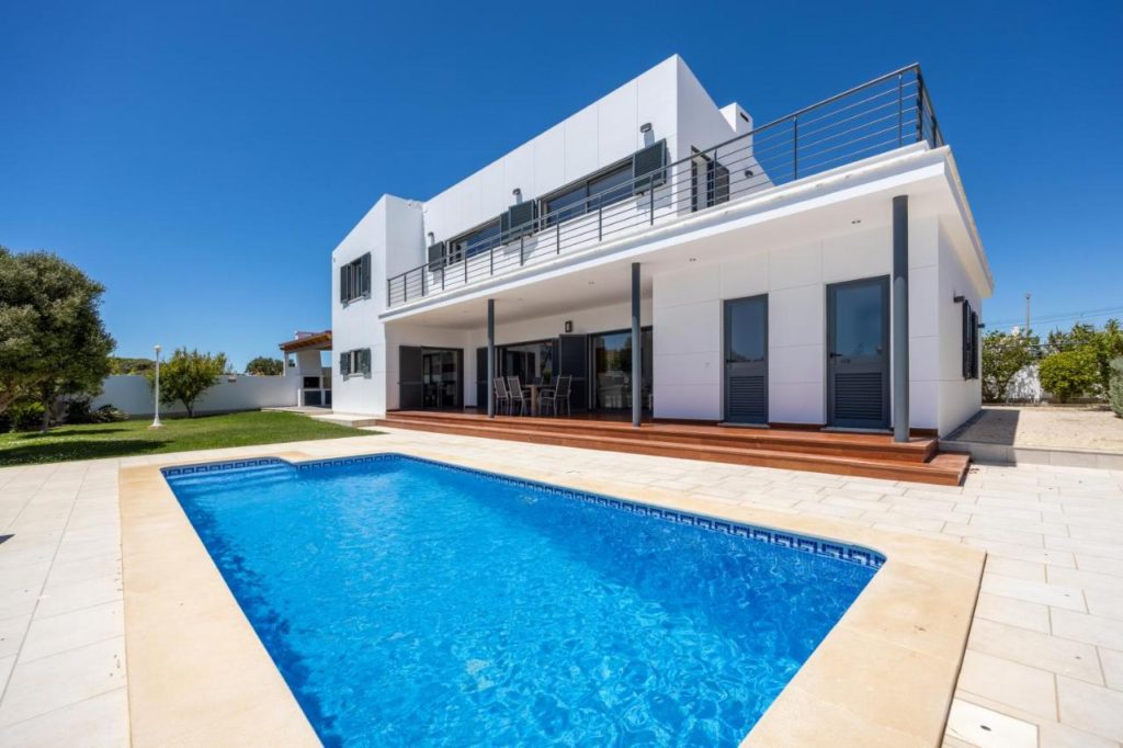 Image of a white villa with a swimming pool in front and a blue sky in the background.